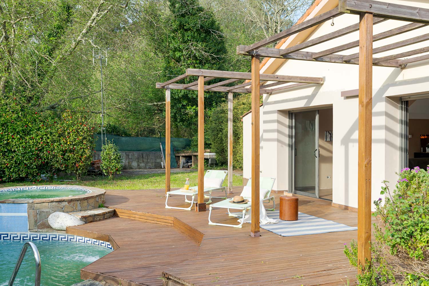 Zona de descanso exterior en una casa unifamiliar, con terraza de madera, pérgola, tumbonas y piscina, ofreciendo un oasis de relajación al aire libre