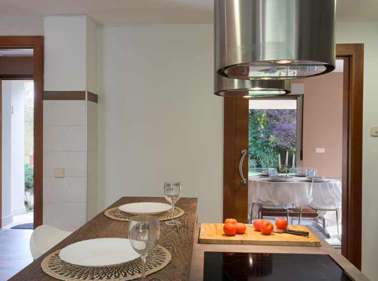 Cocina moderna con una isla de madera, campana extractora de acero, y una vista al comedor exterior a través de una puerta abierta