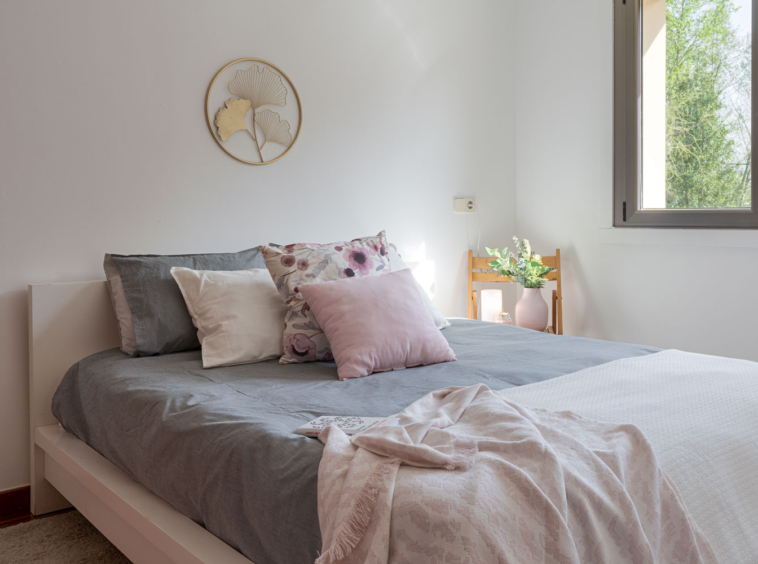Habitación serena con cama amplia, ropa de cama en tonos grises y rosas, y decoración natural, ofreciendo un refugio tranquilo con vista al exterior