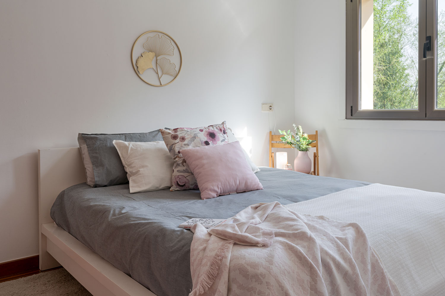Habitación serena con cama amplia, ropa de cama en tonos grises y rosas, y decoración natural, ofreciendo un refugio tranquilo con vista al exterior