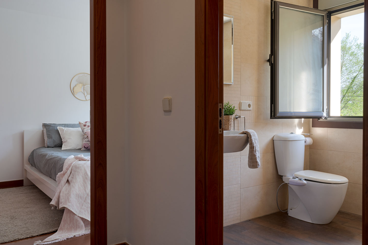 Vista desde el umbral de una puerta que muestra un baño con inodoro y lavamanos y una habitación contigua con cama y decoración suave