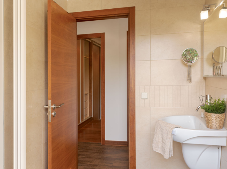 Vista del baño con azulejos beige, espejo moderno, lavabo blanco, y una planta verde, al lado de una puerta abierta que muestra un pasillo de madera