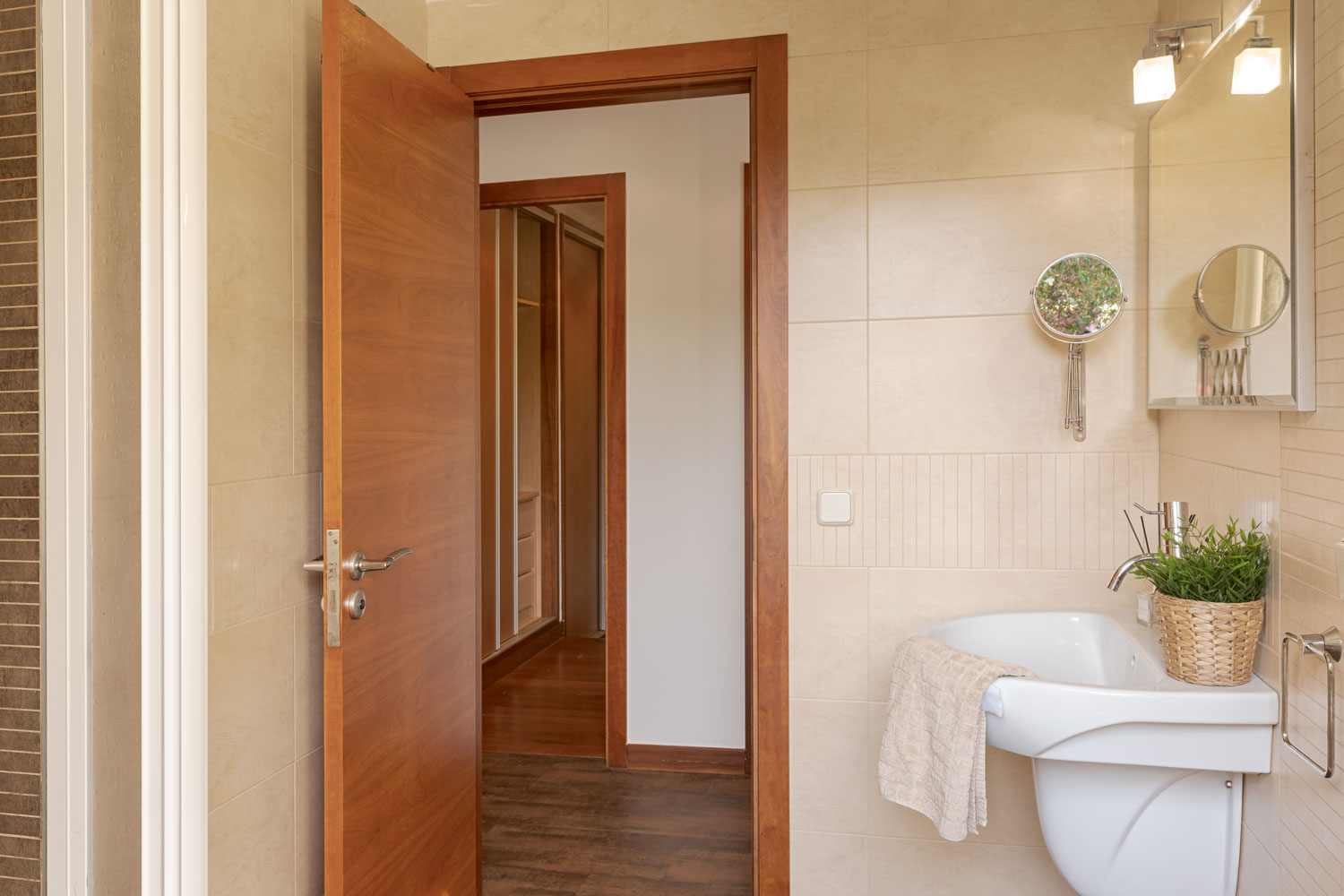 Vista del baño con azulejos beige, espejo moderno, lavabo blanco, y una planta verde, al lado de una puerta abierta que muestra un pasillo de madera