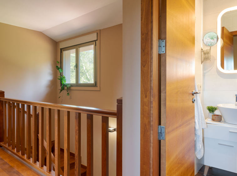 Vista de un pasillo de madera con barandilla que conduce a un baño moderno, ilustrando un hogar contemporáneo con detalles naturales