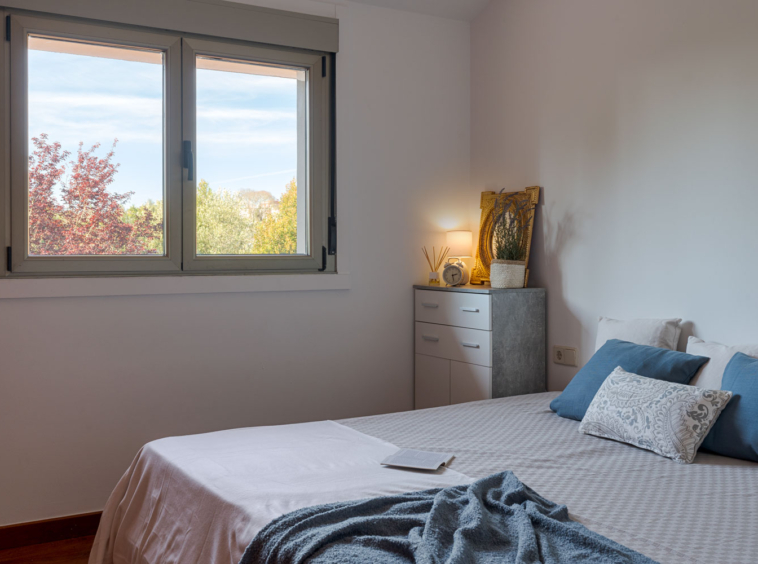 Habitación serena con gran ventana ofreciendo vistas a la vegetación, cama con cojines azules y detalles decorativos que crean un espacio de descanso acogedor