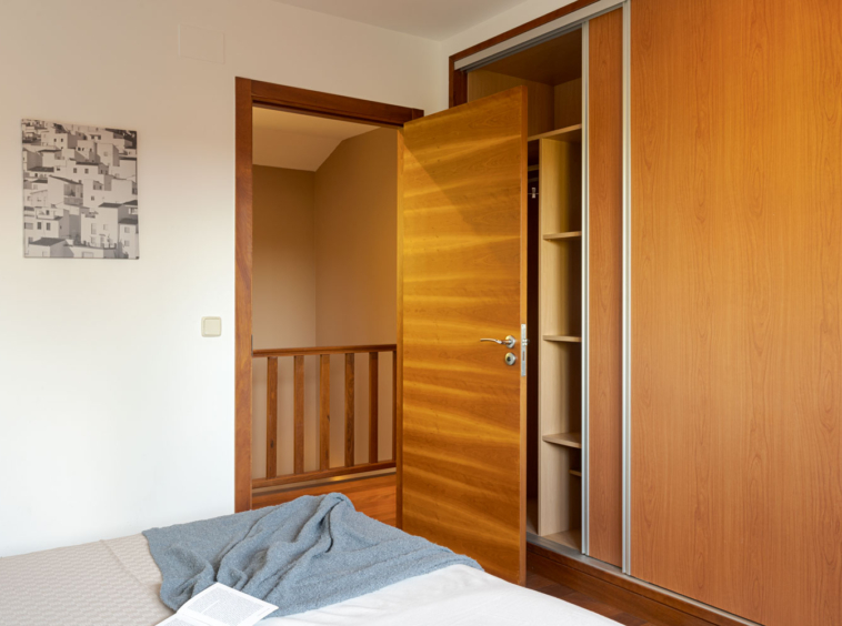 Interior de un dormitorio con una cama y un armario de madera abierto, conectando con un pasillo a través de una puerta de madera.