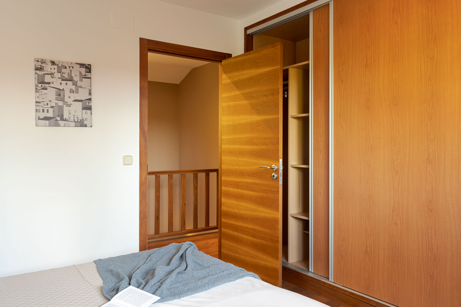 Interior de un dormitorio con una cama y un armario de madera abierto, conectando con un pasillo a través de una puerta de madera.