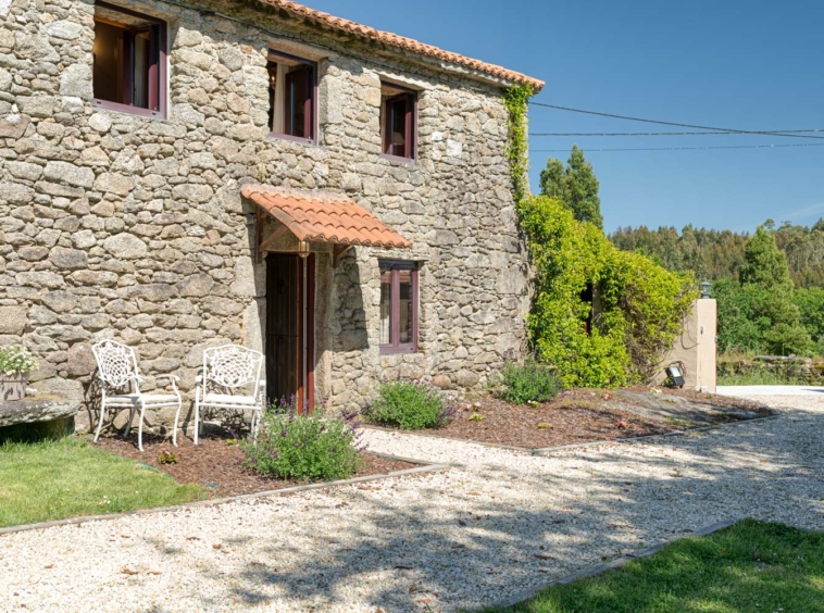 Casa rústica de piedra en venta en Aranga por Inmobiliaria Morando, con dos sillas de metal blancas en la entrada y jardín con flores.