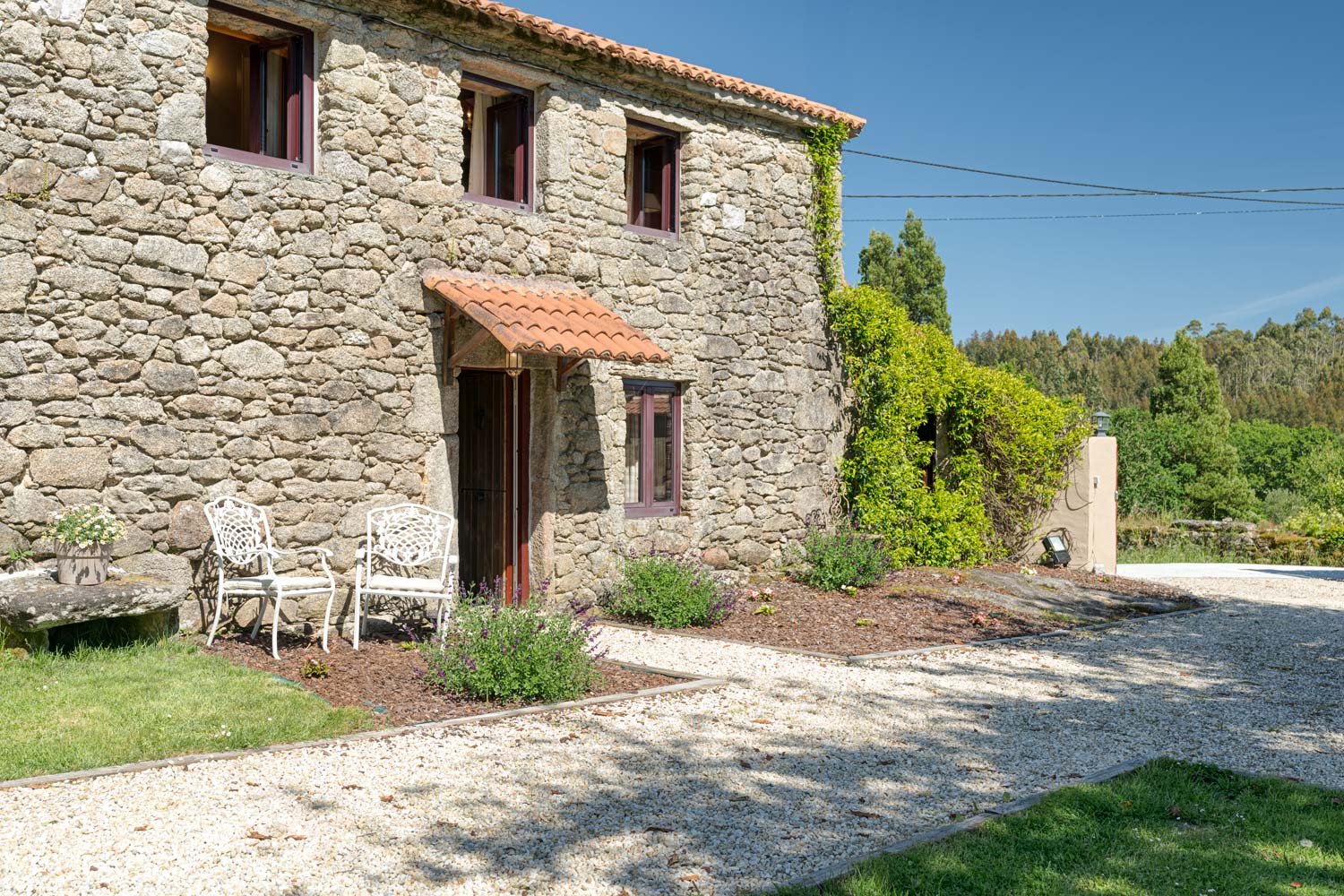 Casa rústica de piedra en venta en Aranga por Inmobiliaria Morando, con dos sillas de metal blancas en la entrada y jardín con flores.