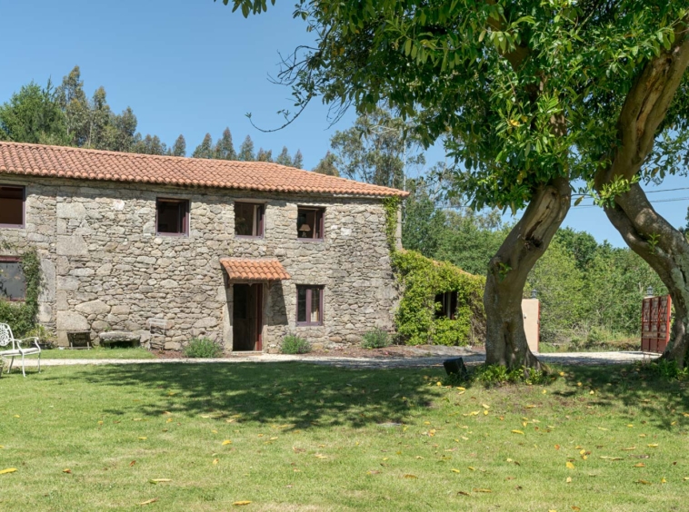 Casa rústica en venta en Aranga por Inmobiliaria Morando, con fachada de piedra, tejado de tejas rojas, y jardín amplio con árboles.