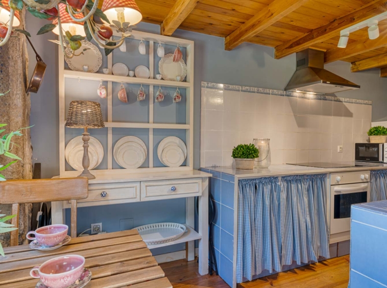 Vista de la cocina con muebles blancos, detalles en azul y un estante de platos decorativos, con iluminación cálida. Casa en venta por Morando en Aranga.