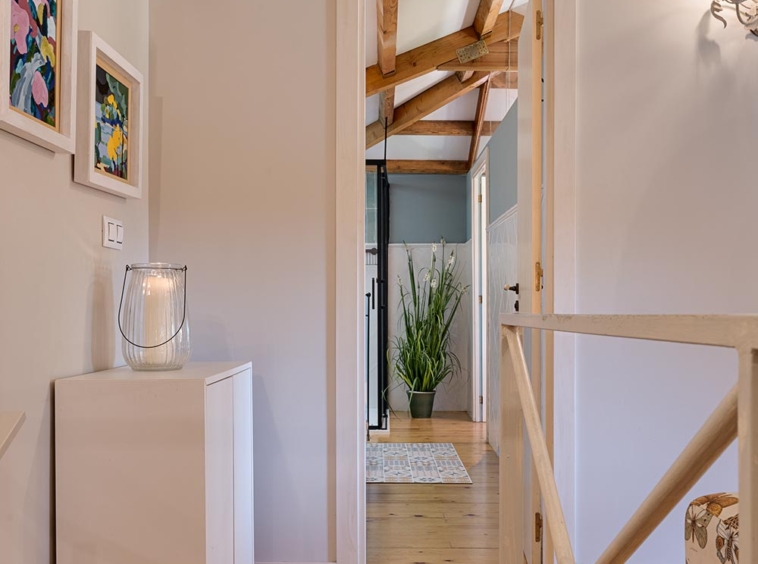 Vista del pasillo con vigas de madera en el techo y piso de madera, que conecta a un baño con plantas decorativas en la distancia. Casa en venta por Morando en Aranga.