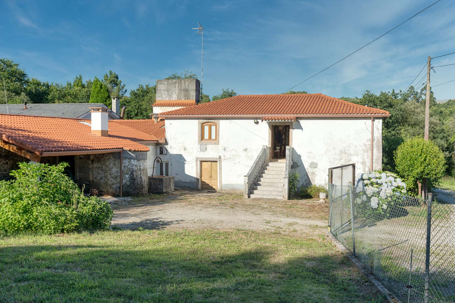 "Casa en venta en Bergondo con tejado de tejas rojas, pozo de agua, jardín y camino de acceso, bajo un cielo azul."