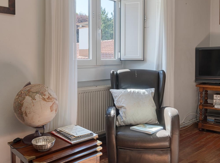 "Rincón de lectura en casa en venta en Bergondo, con sillón de cuero, cojín y mesa auxiliar de madera con un globo terráqueo y libros, junto a una ventana abierta con cortinas blancas."