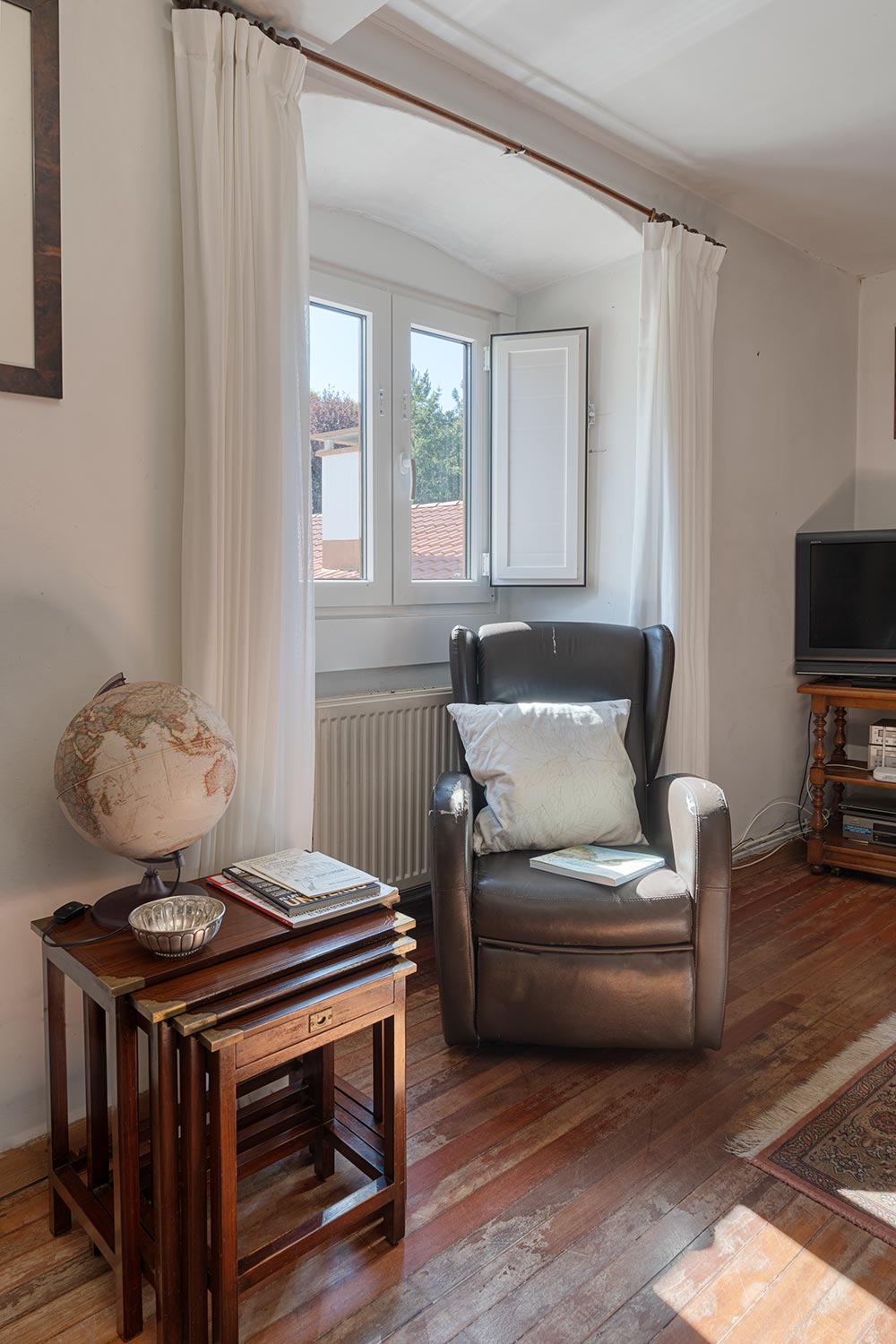 "Rincón de lectura en casa en venta en Bergondo, con sillón de cuero, cojín y mesa auxiliar de madera con un globo terráqueo y libros, junto a una ventana abierta con cortinas blancas."