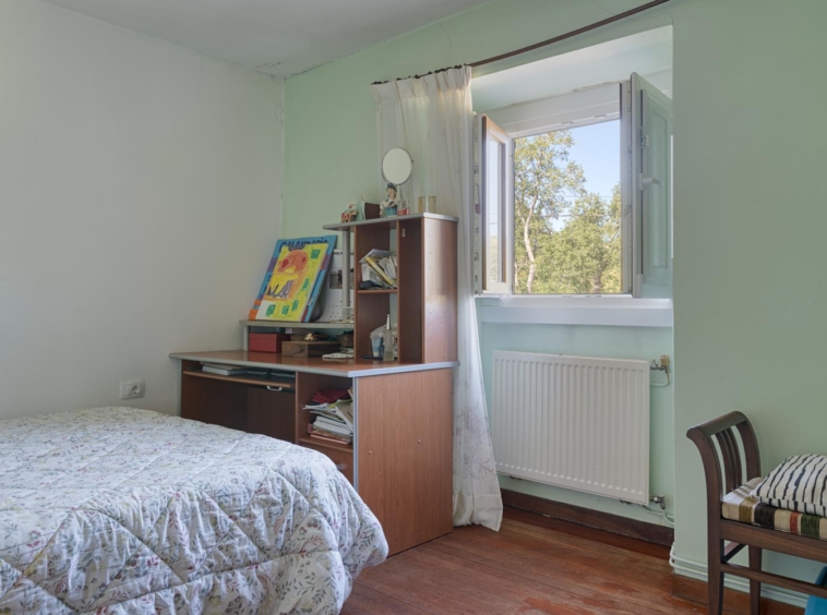 "Pequeño dormitorio en casa en venta en Bergondo, con cama individual, escritorio de madera con estanterías, ventana abierta con vistas al exterior y paredes en color verde claro."