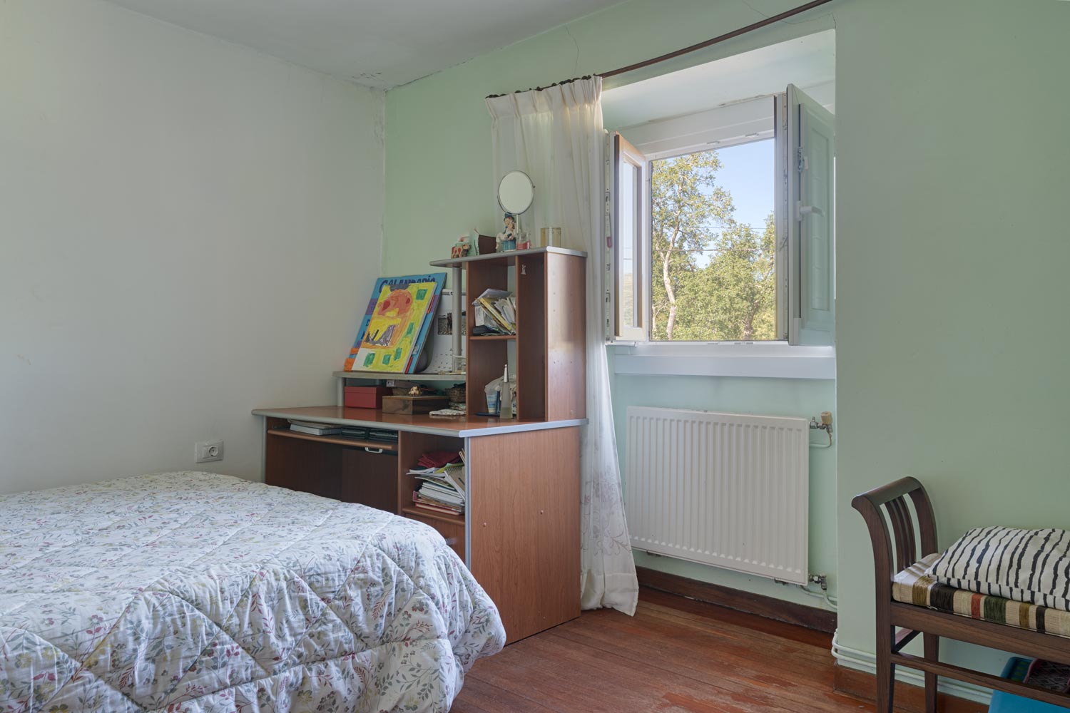 "Pequeño dormitorio en casa en venta en Bergondo, con cama individual, escritorio de madera con estanterías, ventana abierta con vistas al exterior y paredes en color verde claro."