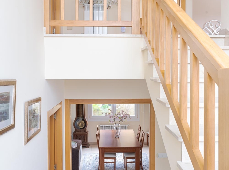 Vista desde una escalera de madera hacia un comedor con mesa de madera y sillas, alfombra clara y detalles decorativos. En venta por Morando Inmobiliaria