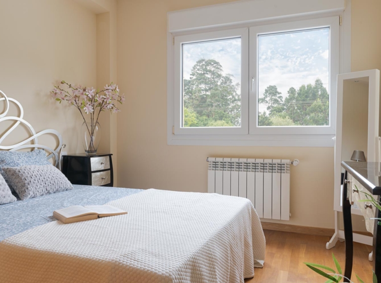 Habitación con cama de diseño elegante, cabecero blanco decorativo, ventana con vista a árboles y radiador debajo. Un libro descansa sobre la cama. En venta por Morando Inmobiliaria.