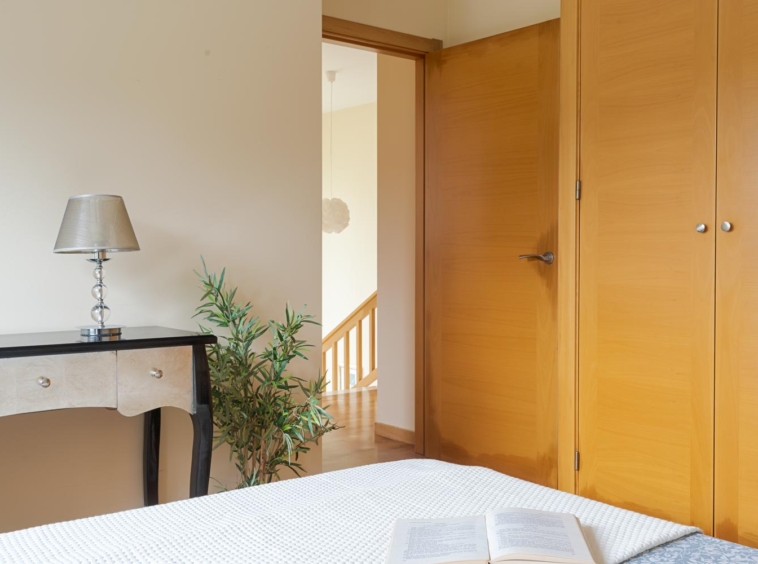 Vista de un dormitorio con una puerta abierta, un armario empotrado de madera y una mesa auxiliar con una lámpara moderna. Un libro abierto reposa sobre la cama. En venta por Morando Inmobiliaria.