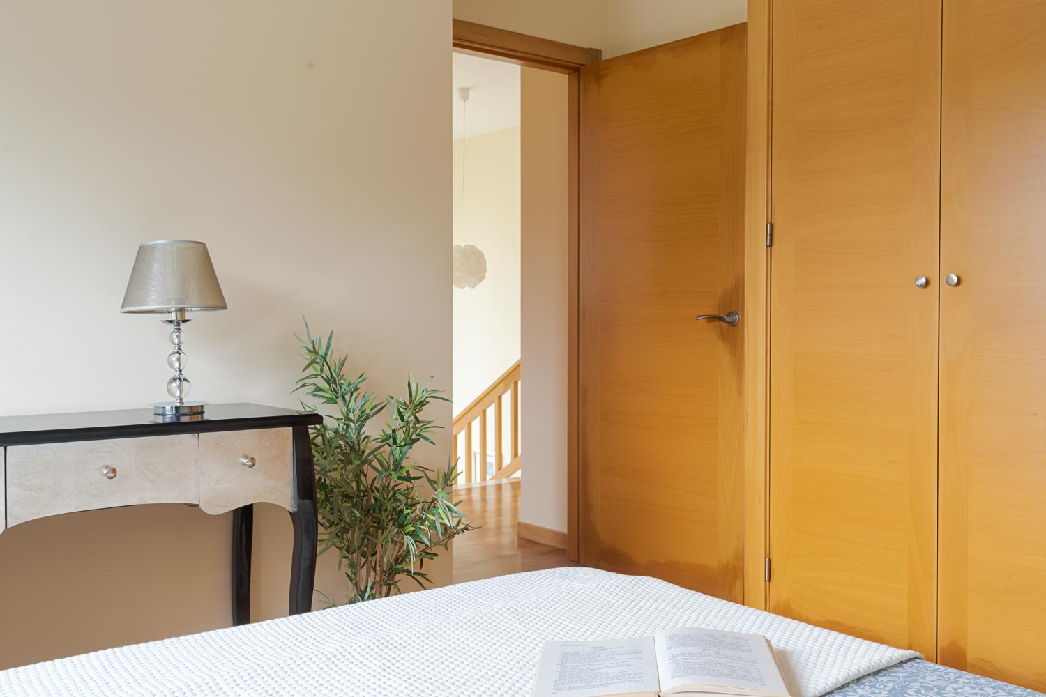 Vista de un dormitorio con una puerta abierta, un armario empotrado de madera y una mesa auxiliar con una lámpara moderna. Un libro abierto reposa sobre la cama. En venta por Morando Inmobiliaria.