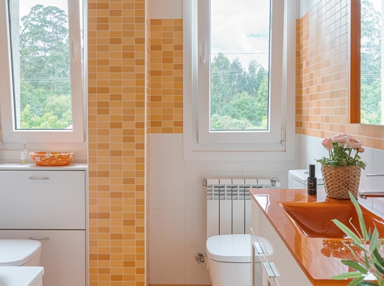 Baño moderno con tonos naranjas y blancos, con dos ventanas grandes que ofrecen vistas a un entorno natural.