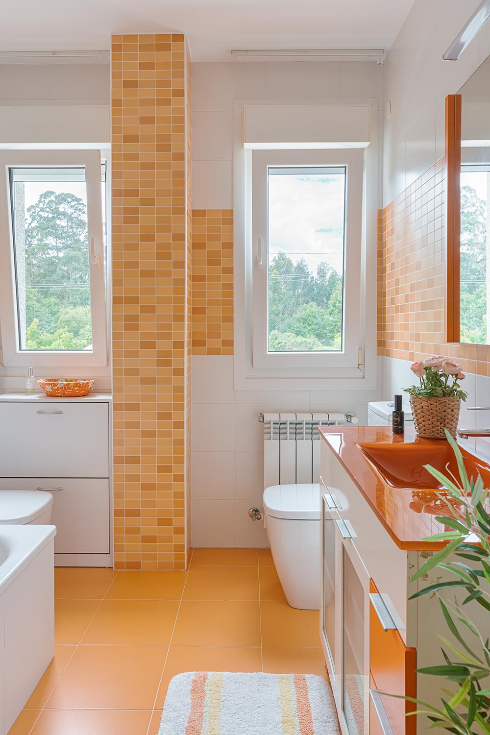 Baño moderno con tonos naranjas y blancos, con dos ventanas grandes que ofrecen vistas a un entorno natural.